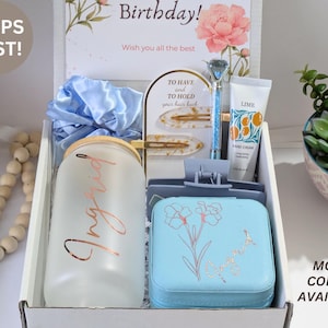 May include: A gift box with various items, including a frosted glass tumbler with "Ingrid" in rose gold, a blue jewelry case with a floral design, and a tube of hand cream. The box also contains a blue scrunchie and hair clips.