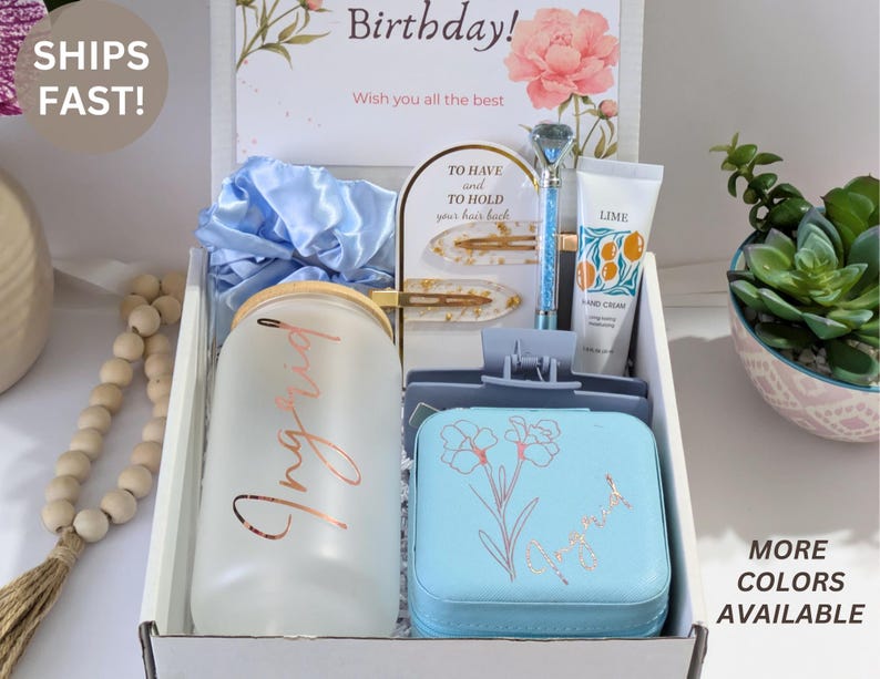 May include: A gift box with various items, including a frosted glass tumbler with "Ingrid" in rose gold, a blue jewelry case with a floral design, and a tube of hand cream. The box also contains a blue scrunchie and hair clips.