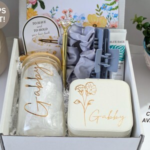 May include: A gift box filled with various items, including a glass jar with "Gabby" in gold lettering, a gold pen, a light blue hair clip, a white jewelry box with floral design and "Gabby" in gold lettering, and a tube of "Blue Sage" cream. The box is white.
