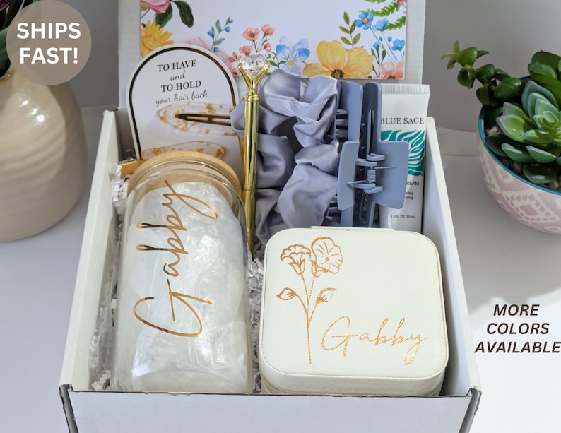May include: A gift box filled with various items, including a glass jar with "Gabby" in gold lettering, a gold pen, a light blue hair clip, a white jewelry box with floral design and "Gabby" in gold lettering, and a tube of "Blue Sage" cream. The box is white.