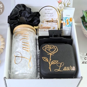 May include: A gift box containing a variety of items. Includes a glass jar with a wooden lid, a black scrunchie, hair clips, a black jewelry box with a gold rose design, and a tube of hand cream. The box is white.