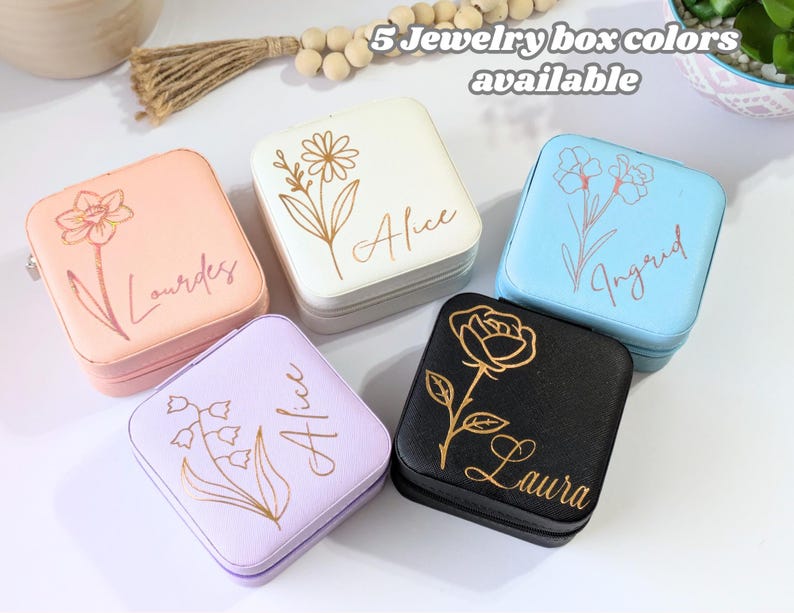 May include: Five square jewelry boxes in various colors: peach, white, light blue, lavender, and black. Each box features a floral design and a name in gold script. The text "5 Jewelry box colors available" is at the top.