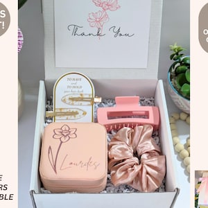 May include: A gift box with hair accessories, including a pink claw clip, scrunchies, and gold barrettes. The box contains a jewelry case with a floral design and the name "Lourdes". A card reads "Thank You".
