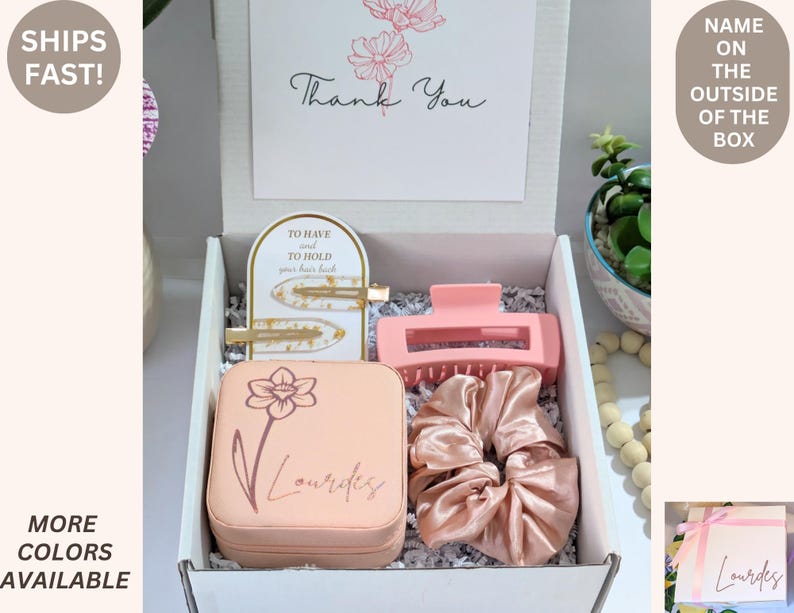 May include: A gift box with hair accessories, including a pink claw clip, scrunchies, and gold barrettes. The box contains a jewelry case with a floral design and the name "Lourdes". A card reads "Thank You".