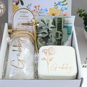 May include: A gift box filled with various items, including a glass jar with "Gabby" in gold script, a pen, hair accessories, a small jewelry box, and a tube of Blue Sage cream. The box is white with floral accents and the text "MORE COLORS AVAILABLE".