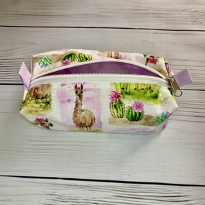 May include: A rectangular, open zipper pouch with a white background featuring a repeating pattern of llamas, cacti, and succulents in a watercolor style. The pouch has a white zipper and a lavender pull tab.