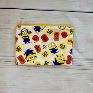 May include: A white zippered pouch with a yellow zipper. The pouch is covered in a pattern of yellow Minions with blue overalls and goggles. The Minions are saying "Bello" and "Kick Me".