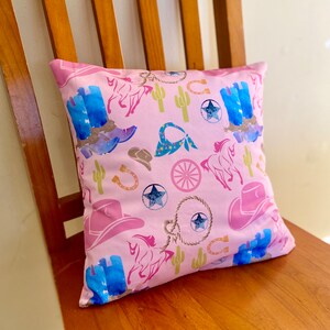 Può includere: Cuscino quadrato rosa con un motivo ripetuto di stivali da cowboy, cappelli, cavalli, cactus, ferri di cavallo e stelle. Gli stivali e le stelle hanno un effetto acquerello. Il cuscino è su una sedia di legno.