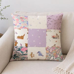 Op de afbeelding: Een vierkant sierkussen met een patchworkontwerp van negen verschillende stoffen vierkanten. De patronen bevatten bloemenprints, paisley-motieven en illustraties van stripfiguren zoals Teigetje en Iejoor in roze, paarse en crèmetinten.