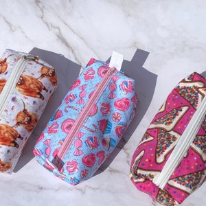 May include: Three zippered pouches with different patterns. One pouch has a blue background with pink and white candy and cupcake designs. Another pouch has a white background with brown Highland cow designs. The third pouch has a pink background with white and colorful sprinkles on a triangle shape.