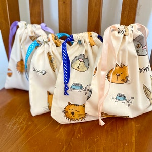 May include: Four drawstring bags with a cream background and a repeating cat-themed pattern. The bags feature illustrations of cat faces, fish bones, and food bowls. Each bag has a different coloured ribbon: purple, blue with white polka dots, blue, and pink.