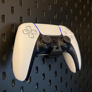 Puede incluir: Un mando de videojuego blanco y negro está montado en una pared de tablero de clavijas negro. El mando tiene una cara blanca y botones y gatillos negros.