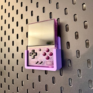 May include: A purple handheld video game console with a screen, buttons, and a purple wall mount. The console is mounted on a pegboard wall.