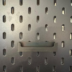 May include: A dark gray pegboard with evenly spaced, oval-shaped holes. A dark gray shelf is inserted into the pegboard. The pegboard is designed for organizing and displaying items.