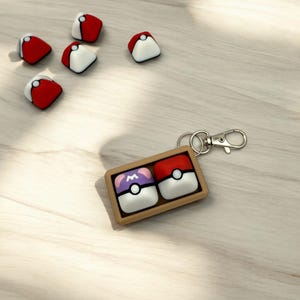 May include: A brown wooden keychain with two Poke Balls. One Poke Ball is red and white, the other is purple and white with an 'M' on it.