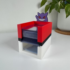 Pode incluir: Um porta-cartões de dois níveis em vermelho e branco, com um detalhe em preto. Uma figura Gengar roxa brilhante está presa no topo. O suporte foi projetado para armazenar cartas colecionáveis.