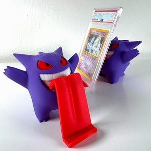 May include: A purple Gengar figure with red eyes and a red tongue, holding a graded Pokémon card in a clear case. The card features a Mewtwo illustration. Another Gengar figure is visible in the background.