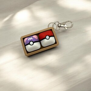 May include: A brown wooden box with a silver keychain attached. The box contains two small, square, colorful Poke Balls. One Poke Ball is red with a white circle and black dot. The other Poke Ball is purple with a white circle and black dot. The Poke Balls have a white rim.