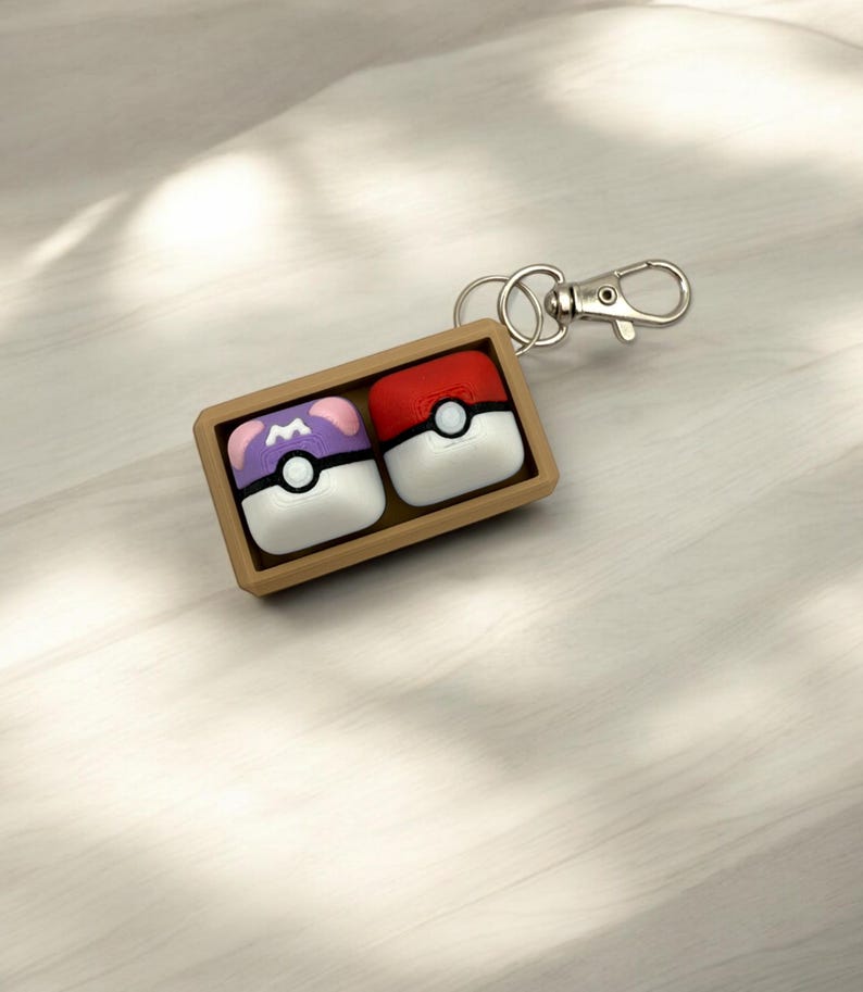May include: A brown wooden box with a silver keychain attached. The box contains two small, square, colorful Poke Balls. One Poke Ball is red with a white circle and black dot. The other Poke Ball is purple with a white circle and black dot. The Poke Balls have a white rim.
