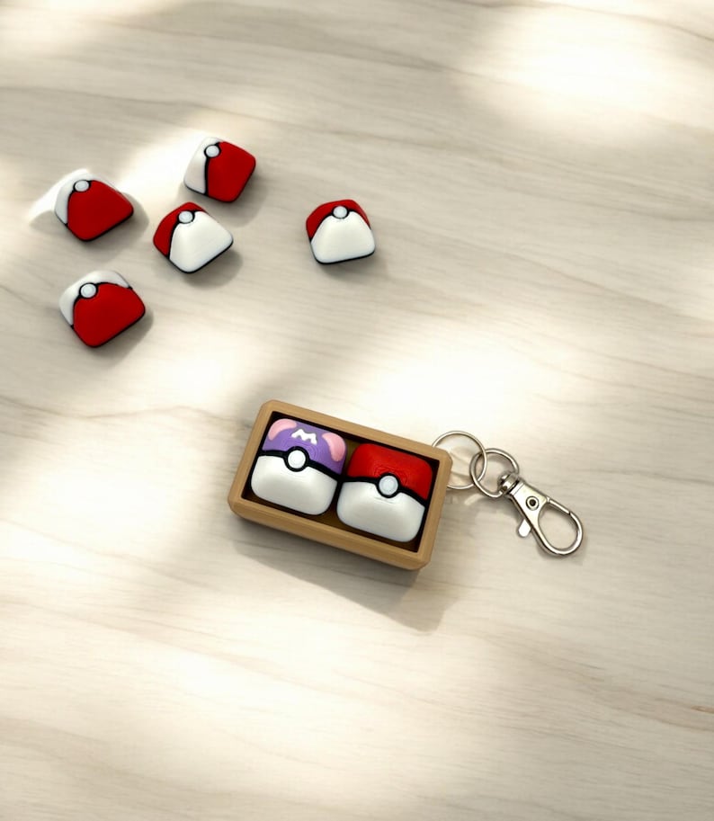 May include: A keychain with a wooden box containing two Poke Balls. One Poke Ball is red and white, the other is purple and white. The keychain has a silver metal clasp.