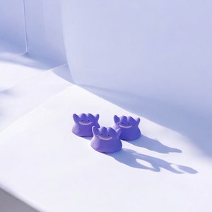 May include: Three small, lavender-colored Ditto figurines are arranged on a white surface. Each figure has a smiling face and a blob-like form. The background is a gradient of white and light purple.