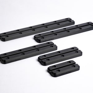 Puede incluir: Un conjunto de piezas de hardware negras y rectangulares con múltiples agujeros, dispuestas sobre una superficie blanca. Las piezas varían en tamaño, algunas más largas y otras más cortas. El texto "Leather&Dirt" es visible en algunas piezas.
