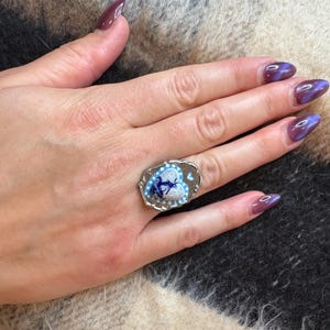 May include: A silver ring with an oval face. The ring features a heart-shaped design with a blue and white floral pattern and a central image of a person. The ring is on a hand with purple nail varnish.
