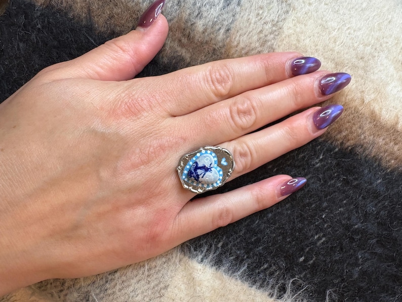 May include: A silver ring with an oval face. The ring features a heart-shaped design with a blue and white floral pattern and a central image of a person. The ring is on a hand with purple nail varnish.