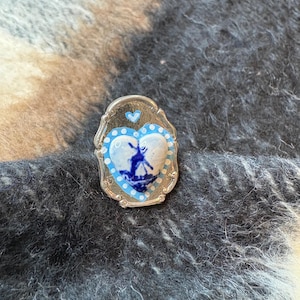 May include: A silver-coloured pin with a heart-shaped blue and white design. The heart features a windmill scene and is surrounded by a blue border and white dots. A small blue heart is at the top of the pin.