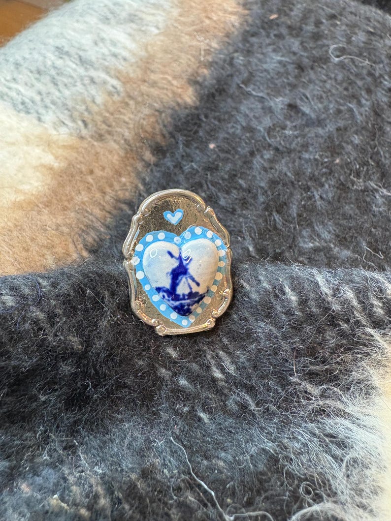 May include: A silver-coloured pin with a heart-shaped blue and white design. The heart features a windmill scene and is surrounded by a blue border and white dots. A small blue heart is at the top of the pin.