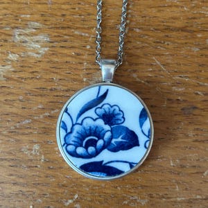 May include: A circular pendant necklace featuring a white ceramic inlay with a blue floral pattern. The pendant is set in a silver-toned metal frame and hangs from a matching silver-toned metal chain link necklace.
