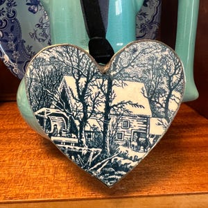 May include: A heart-shaped ceramic ornament featuring a blue and white winter scene of a snow-covered house and bare trees. The ornament is edged in silver metal and hangs from a black velvet ribbon, showcasing a detailed landscape illustration.