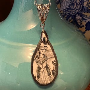 May include: Silver-toned pendant necklace featuring a teardrop-shaped pendant with a black and white illustration of a woman in period clothing. The pendant hangs from a delicate silver chain. The background is a turquoise vase.