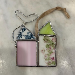 May include: A stained glass house ornament with a pink panel, floral design, and blue patterned roof. The other house has a green roof and floral design. A silver wire heart and twine loop are attached for hanging.
