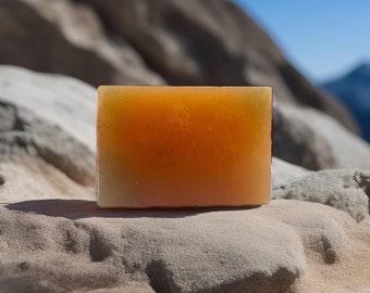 May include: A rectangular bar of orange soap with a slight green tint, sitting on a light brown, sandy surface.
