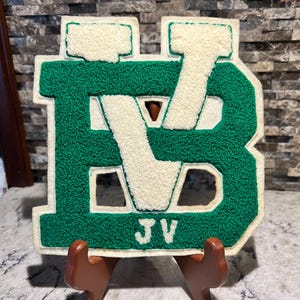 May include: A green and cream-colored varsity letter patch with the letters "BV" and the initials "JV" at the bottom. The patch is displayed on a brown stand against a textured background.