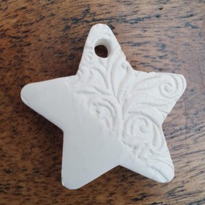 May include: A white star-shaped decoration with a hole for hanging. One side of the star has an embossed floral pattern. The ornament is made of a matte, off-white material. The star is approximately 7 cm wide.