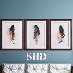 May include: Three framed prints of watercolour paintings of black feathers with a light pink and orange background. The prints are hanging on a blue wall above a white sofa.