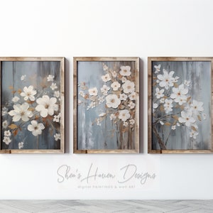 May include: Set of three framed canvas prints featuring a floral design with white flowers and brown branches on a blue and grey background. The frames are a light brown wood.
