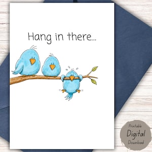 Può includere: Un biglietto d'auguri con un'illustrazione di tre uccellini azzurri con il becco arancione appollaiati su un ramo marrone. Due uccelli sono seduti mentre il terzo è appeso al ramo. Il testo Hang in there è stampato sopra l'immagine.