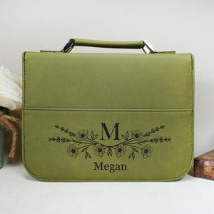 May include: A green faux leather Bible cover with a floral design and the initial "M" and the name "Megan" engraved on the front.