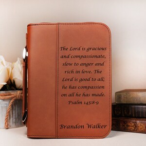 May include: A brown leather Bible cover with a zipper closure and a silver handle. The cover is personalized with the text "The Lord is gracious and compassionate, slow to anger and rich in love. The Lord is good to all; he has compassion on all he has made. Psalm 145:8-9" and the name "Brandon Walker".