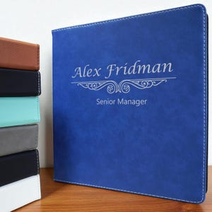 May include: A blue leather binder with white stitching and the name "Alex Fridman" and "Senior Manager" in silver lettering. A stack of binders in various colors is visible to the left.