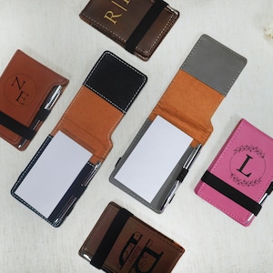 May include: Six different personalized leatherette notebooks with a pen holder and a notepad. The notebooks are in various colors, including brown, black, gray, pink, and orange. Each notebook has a different monogram or design, including the letters "NE", "L", and "R".