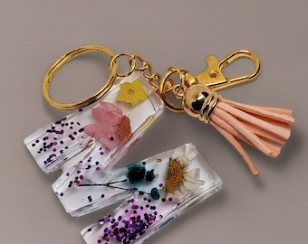 May include: Keyrings with clear resin rectangles, each containing dried flowers and purple glitter. The keyrings have gold-coloured hardware, including a keyring, clasps, and a tassel. The tassel is a light peach colour.