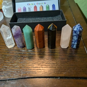 May include: Set of seven chakra crystals in a black box. The crystals are arranged in a row from left to right: clear, amethyst, green, red, black, pink, and blue.
