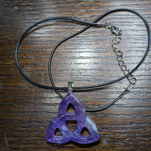 May include: A purple amethyst pendant in a Celtic knot design, suspended from a black cord necklace with a silver-tone clasp. The pendant is a deep violet color and has three interlocking sections.