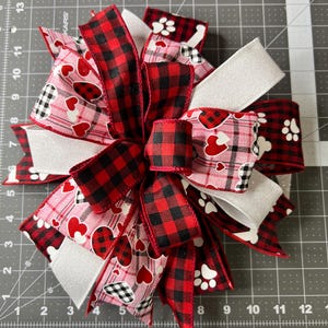 May include: A red and black buffalo plaid ribbon, a white ribbon with a pink and white heart and paw print pattern, and a white ribbon with glitter are tied together in a bow.