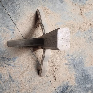 May include: A close-up of a wooden table leg with a decorative design. The leg is made of dark wood and has a smooth finish. The leg is resting on a concrete floor covered in sawdust.