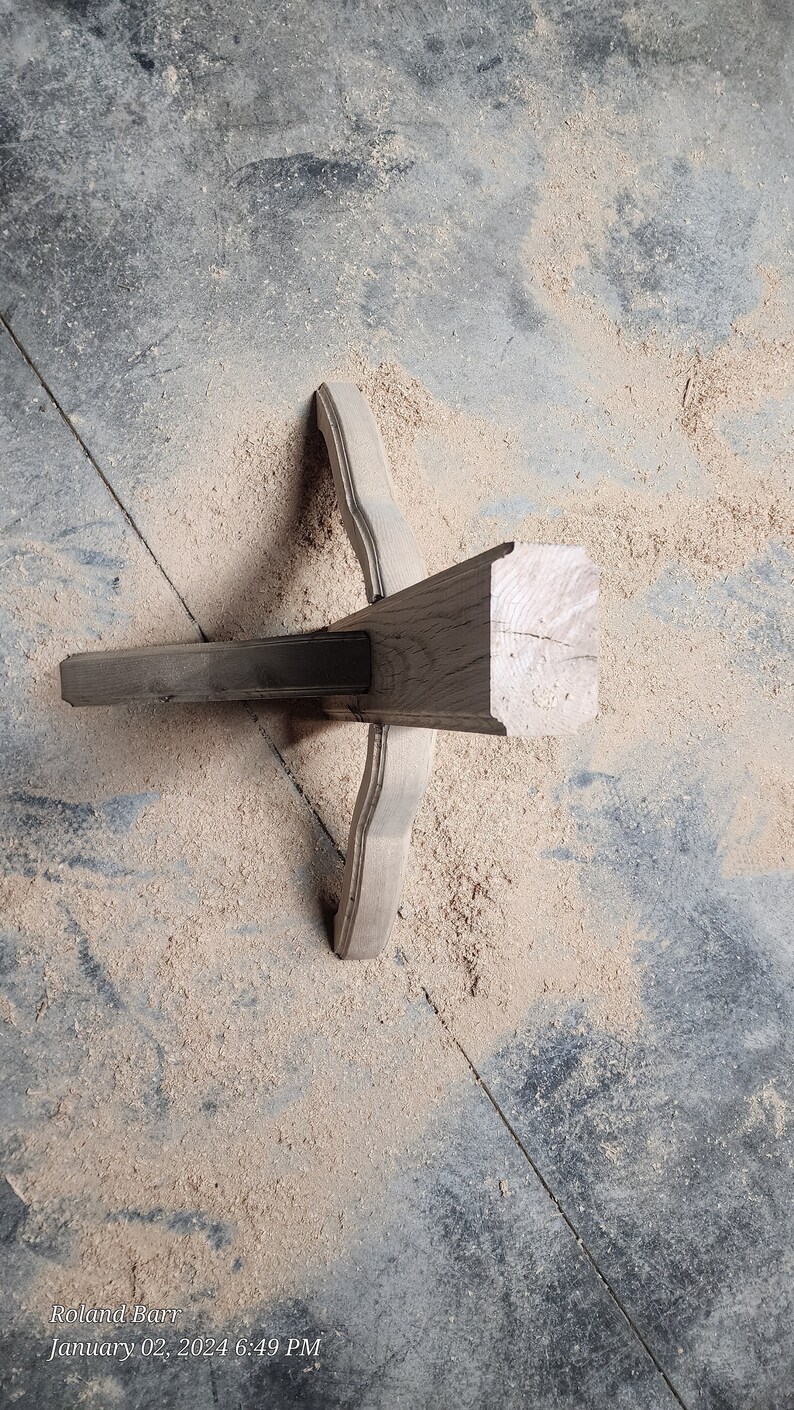 May include: A close-up of a wooden table leg with a decorative design. The leg is made of dark wood and has a smooth finish. The leg is resting on a concrete floor covered in sawdust.